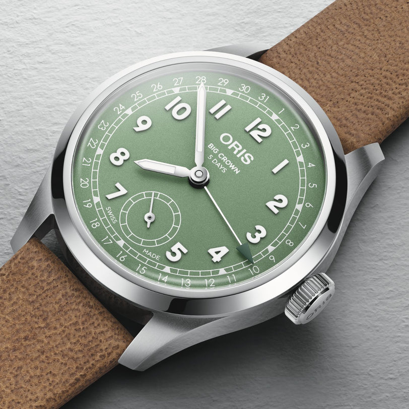 The Oris Big Crown Pointer Date Calibre 403 features a green dial with white numerals, small seconds subdial at 6 o’clock, and a brown leather strap, exemplifying Swiss watch design as it rests on a light gray surface.