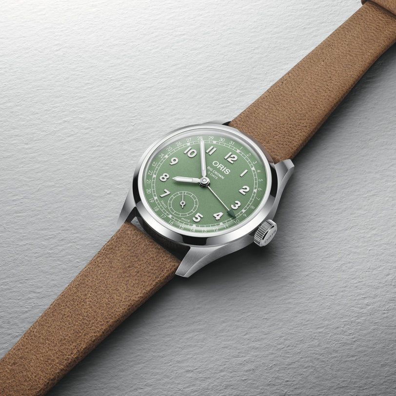 The Oris Big Crown Pointer Date Calibre 403, featuring a light green dial with white numerals and a brown leather strap, exemplifies classic Swiss watch design and is shown resting on a textured gray surface.