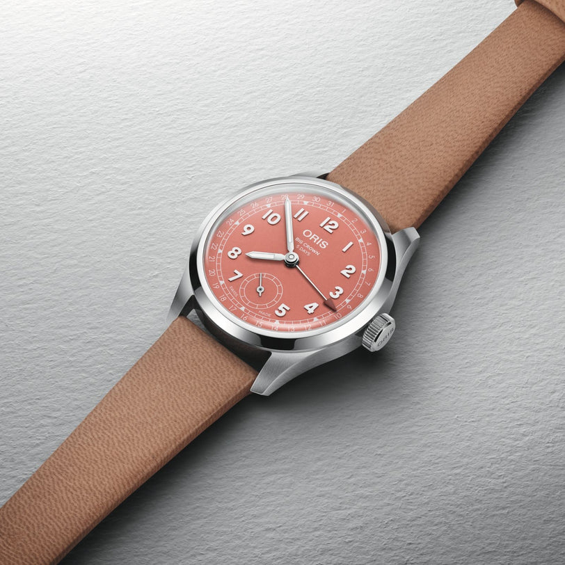 The Oris Big Crown Pointer Date Calibre 403 features a terracotta dial with white numerals and a tan leather strap, displayed on a light gray textured surface.