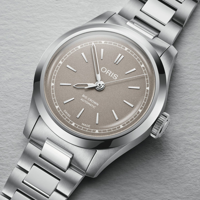 The Oris Big Crown is a Swiss Made automatic watch with a stainless steel case, beige dial, silver markers and hands, date at 6 o’clock, and a metal bracelet.