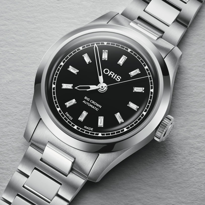 An Oris Big Crown Diamonds wristwatch with a black dial and silver hour markers on a metal band sits on a gray surface.