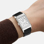 A Jaeger-LeCoultre Reverso Tribute Duoface Small Seconds, a rectangular steel wristwatch with a white dial, small seconds feature, and black leather strap, is worn on the wrist of someone in a brown sweater.