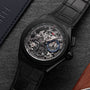 The Zenith Defy El Primero 21, a black luxury skeleton watch with visible mechanical movement, high-frequency chronograph, black leather strap, and blue accents, rests on a dark textured surface near leather, exuding urban sophistication.