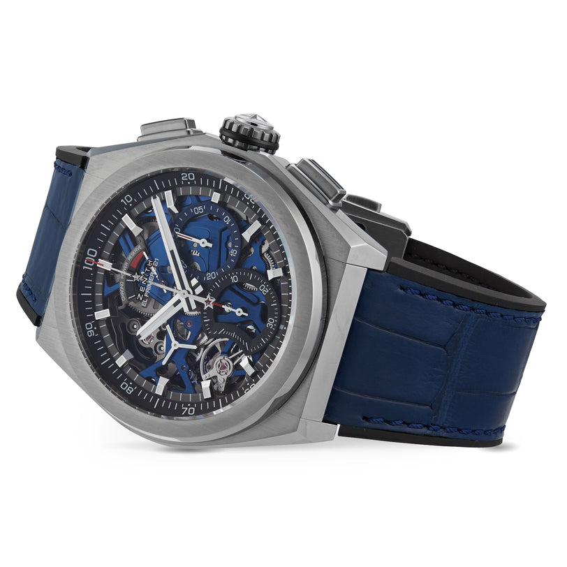 The Zenith Defy El Primero 21 features a titanium DEFY case, blue openworked dial, chronograph functions, and a blue leather strap, shown at an angle.