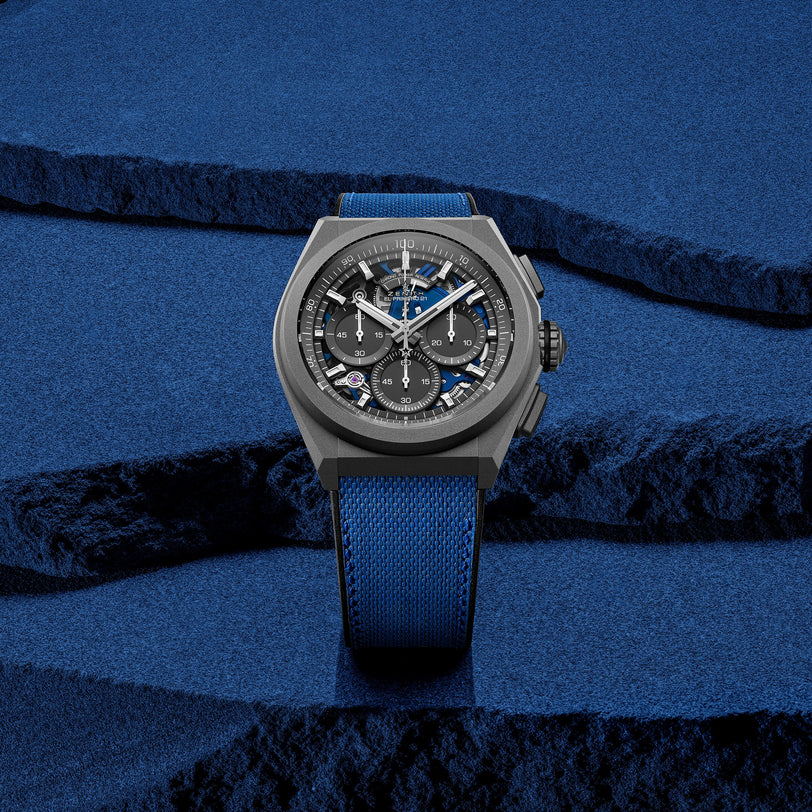 The Zenith Defy 21 Ultrablue wristwatch, featuring a blue strap and black dial, is showcased on a textured blue background with layered surfaces, highlighting the brand’s heritage.