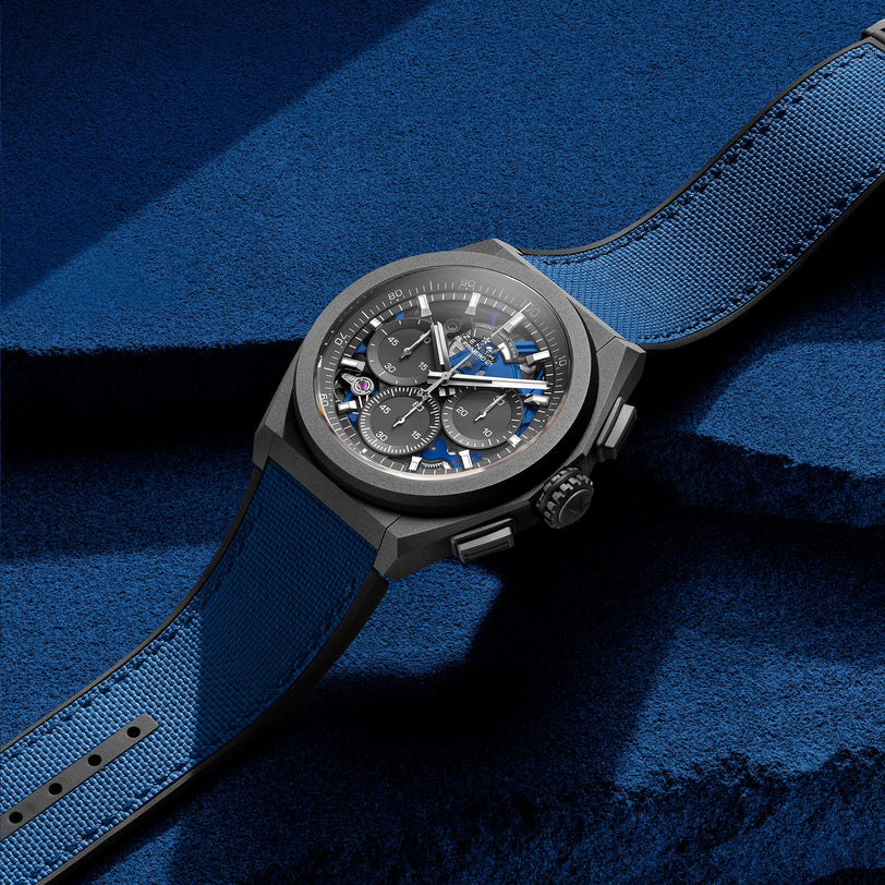 The Zenith Defy 21 Ultrablue chronograph, featuring a fabric strap in black and blue, is displayed on a textured blue surface, showcasing the brands heritage.
