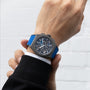A person in a black jacket showcases the Zenith Defy 21 Ultrablue wristwatch, featuring a blue strap and an intricate dark analog face, set against a light, textured background.
