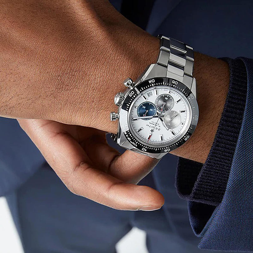 A person in a dark blue suit wearing a Zenith Chronomaster Sport wristwatch with a silver case and a white and blue dial.