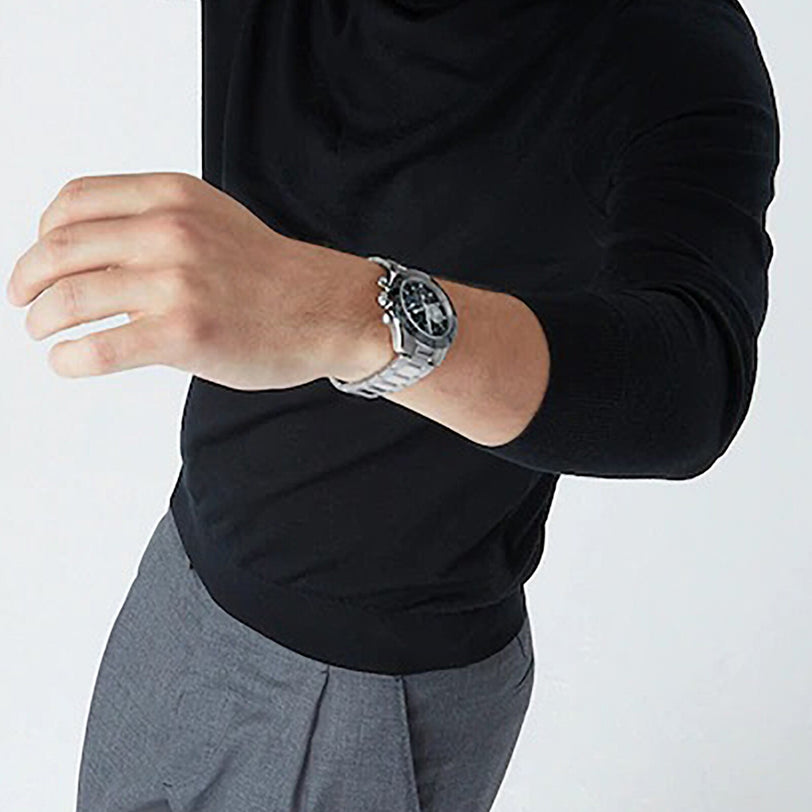 A person in a black long-sleeve shirt and gray pants is shown from the shoulders down, wearing a silver Zenith Chronomaster Sport chronograph on their raised left arm.