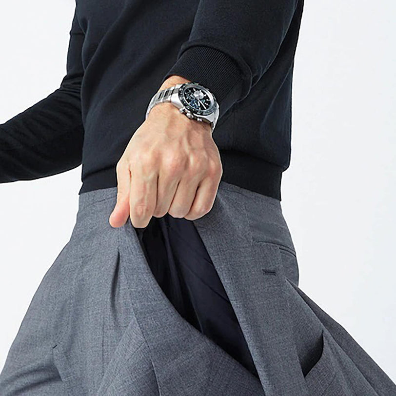 A person in a black long-sleeve shirt and Zenith Chronomaster Sport watch pulls open the side of their gray suit pants, revealing the inner lining.