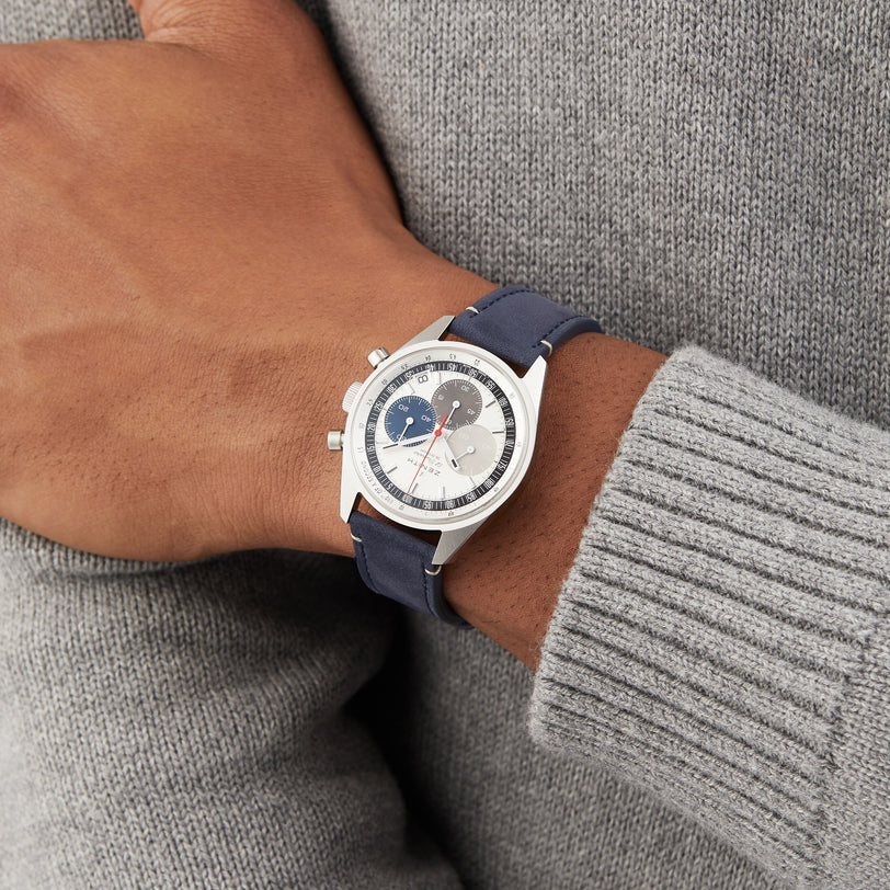 A person in a gray sweater showcases the Zenith Chronomaster EP Original, featuring a silver case, blue strap, and a round white tricolour dial with blue and gray subdials.