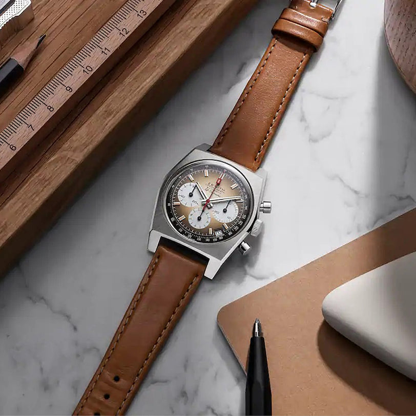 The Zenith Chronomaster Revival El Primero A384 wristwatch with a brown leather strap rests on a marble desk alongside a ruler, notebook, and pen.