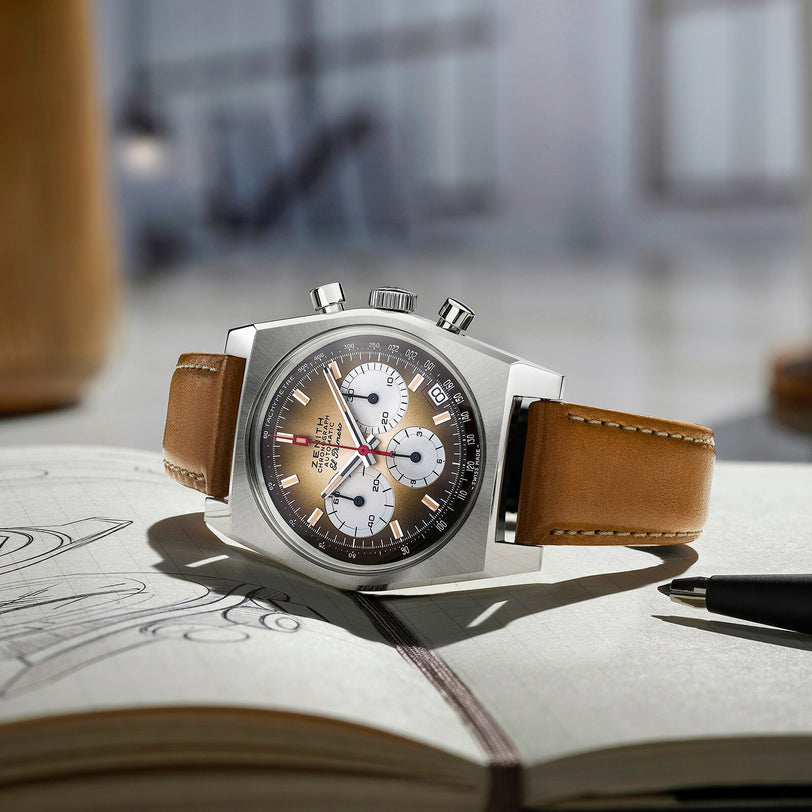 A Zenith Chronomaster Revival El Primero A384 with a silver case, high-frequency chronograph, and brown leather strap rests on an open notebook with sketches, beside a black pen in a softly lit room.