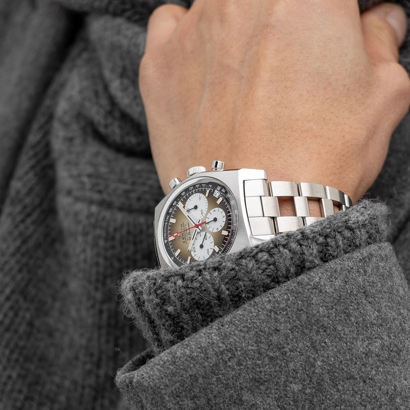 A person wearing a Zenith Chronomaster Revival El Primero A384 silver chronograph wristwatch with a metal band, dressed in a grey textured coat or sweater.