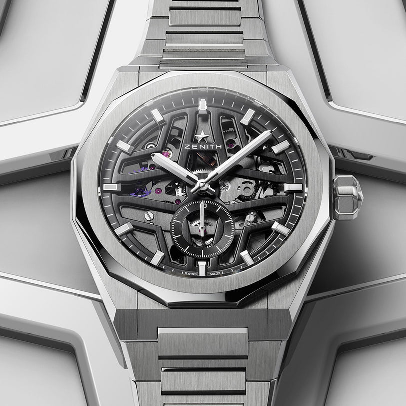 A Zenith Defy Skyline Skeleton watch with a stainless steel bracelet and skeleton dial, showcasing the El Primero 3620 movement, is placed on a geometric metallic surface.