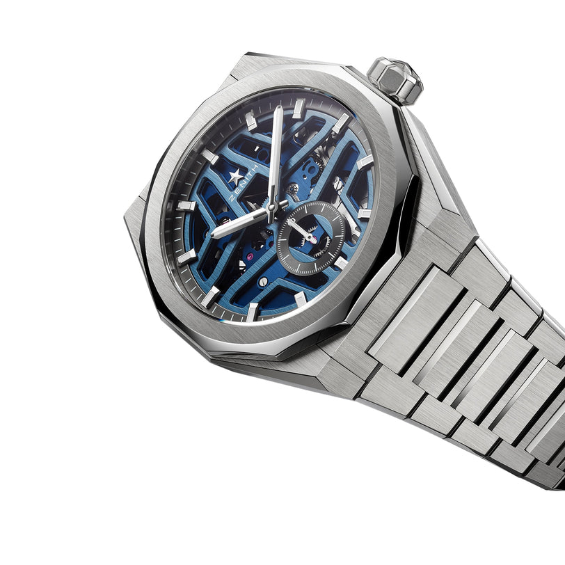 The Zenith Defy Skyline Skeleton is a luxury silver wristwatch featuring an urban blue skeleton dial with El Primero movement, white hands, a metal bracelet, and an octagonal bezel set against a white background.