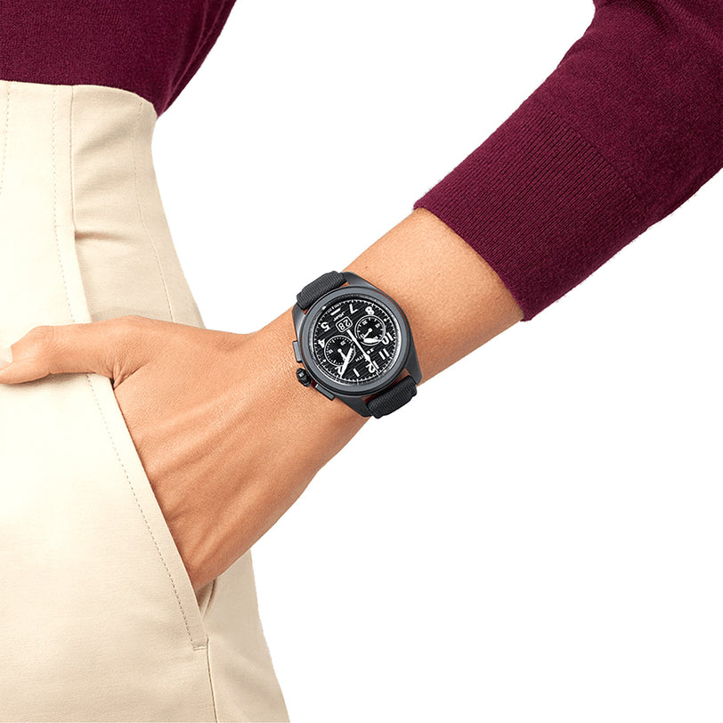 A person in a burgundy long-sleeve top and beige pants wears the Zenith Pilot Big Date Flyback, a stylish black ceramic case watch, with one hand in their pocket against a white background.
