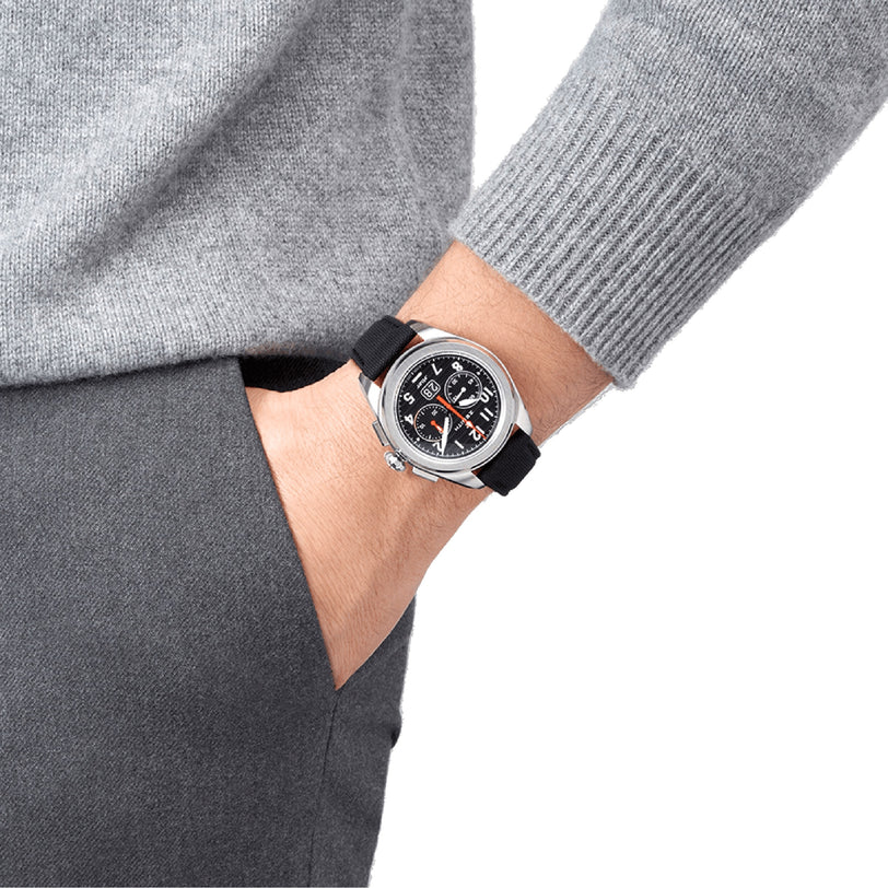 A person in a gray sweater and pants has their hand in their pocket, showcasing the Zenith Pilot Big Date Flyback—a black and silver pilot watch with an analog face and sleek black ceramic case.