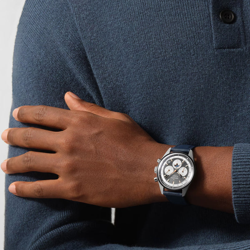 A person in a blue long-sleeve shirt crosses their arms, showing off a Zenith Chronomaster Original chronograph wristwatch with a black and silver dial and dark strap on their wrist.