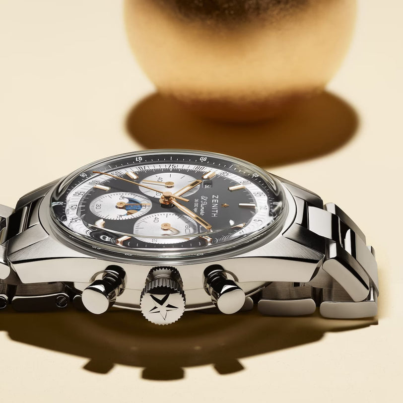 A Zenith Chronomaster Original stainless steel chronograph wristwatch with a black bezel, silver and white subdials, and a metal bracelet is shown on a light surface with a blurred gold object in the background.