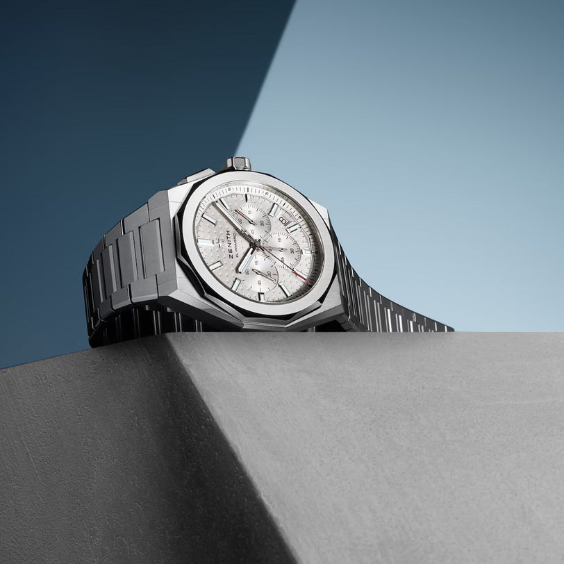 A Zenith Defy Skyline Chronograph wristwatch with a textured dial and interchangeable steel bracelet is displayed on a gray angled surface against a blue background.