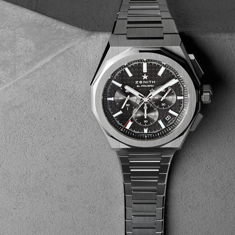 A Zenith Defy Skyline Chronograph watch with a stainless steel bracelet rests on a gray textured surface.
