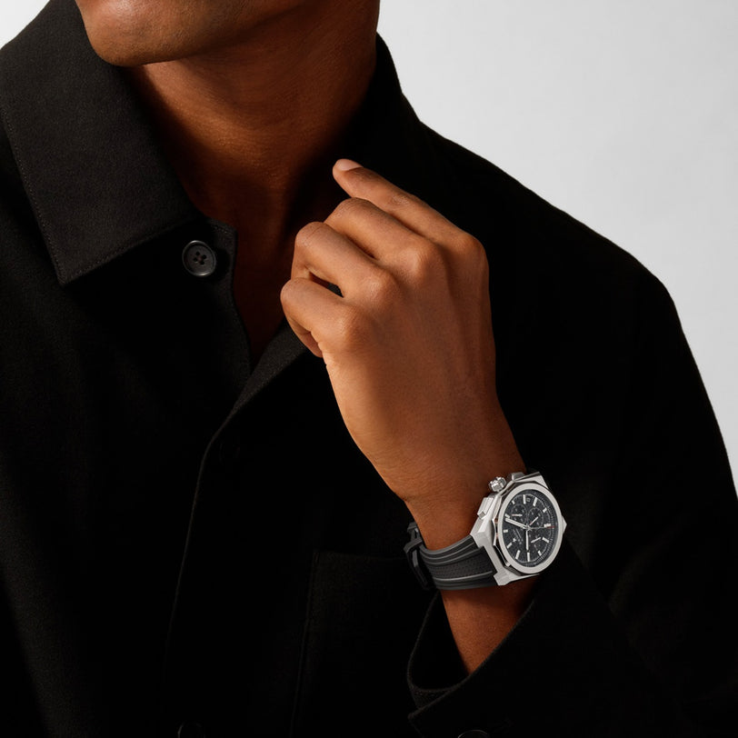 Someone raises their left hand toward their collar, wearing a black shirt and the Zenith Defy Skyline Chronograph, a silver wristwatch featuring an interchangeable strap.
