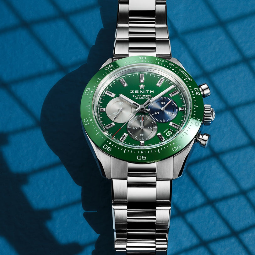 A Zenith Chronomaster Sport watch with a green ceramic bezel, green dial, three sub-dials, and a silver metal bracelet is shown on a blue surface with grid-like shadows.