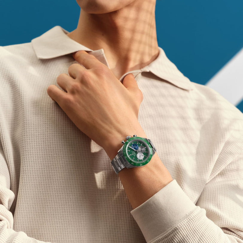 A person in a light beige textured shirt showcases the Zenith Chronomaster Sport silver wristwatch featuring a green ceramic bezel and El Primero 3600 movement, set against a blue background.