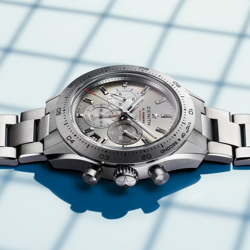A Zenith Chronomaster Sport chronograph watch with a silver dial, triple sub-dials, and a metal bracelet is shown resting on a surface featuring a blue grid shadow pattern.