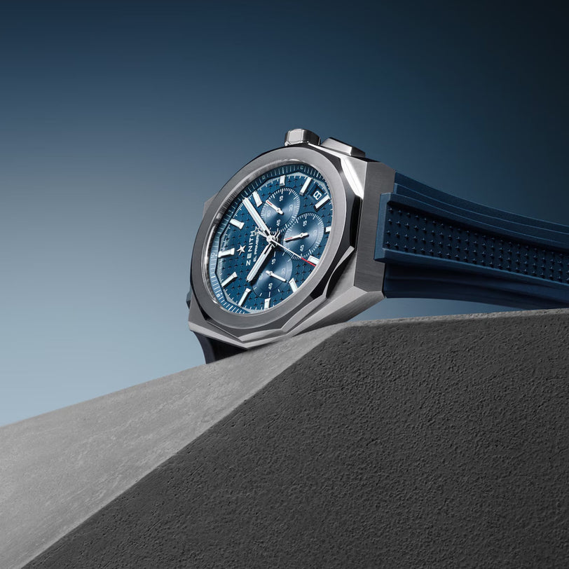A Zenith Defy Skyline Chronograph wristwatch with a blue dial and rubber strap, featuring the El Primero 3600 movement and an interchangeable bracelet, is showcased on a gray angular surface with a gradient blue background.