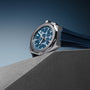 A Zenith Defy Skyline Chronograph wristwatch with a blue dial and rubber strap, featuring the El Primero 3600 movement and an interchangeable bracelet, is showcased on a gray angular surface with a gradient blue background.