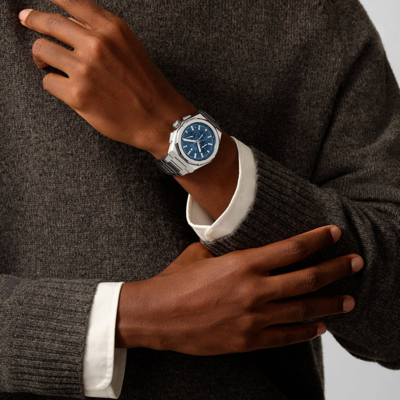 A person in a gray sweater and white shirt showcases a Zenith Defy Skyline Chronograph with a striking blue dial and interchangeable bracelet on their left wrist.