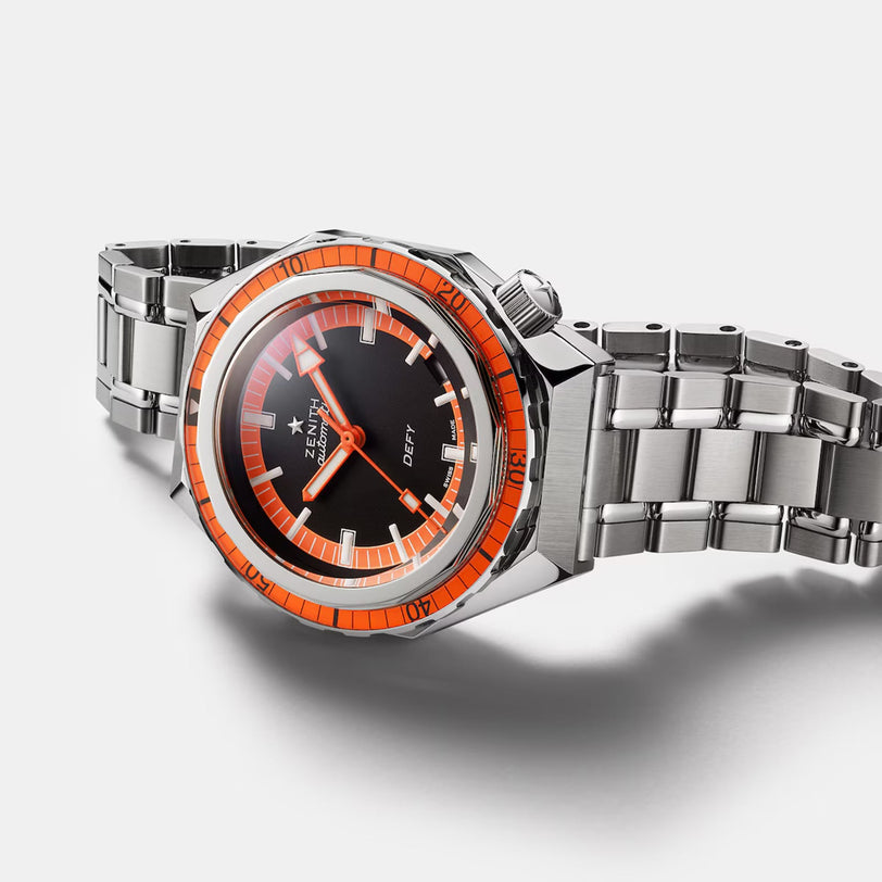 The Zenith Defy Revival is a stainless steel diver’s watch with an orange unidirectional bezel, black dial, silver bracelet, and Elite 670 automatic movement. It features the Zenith logo and Defy branding.