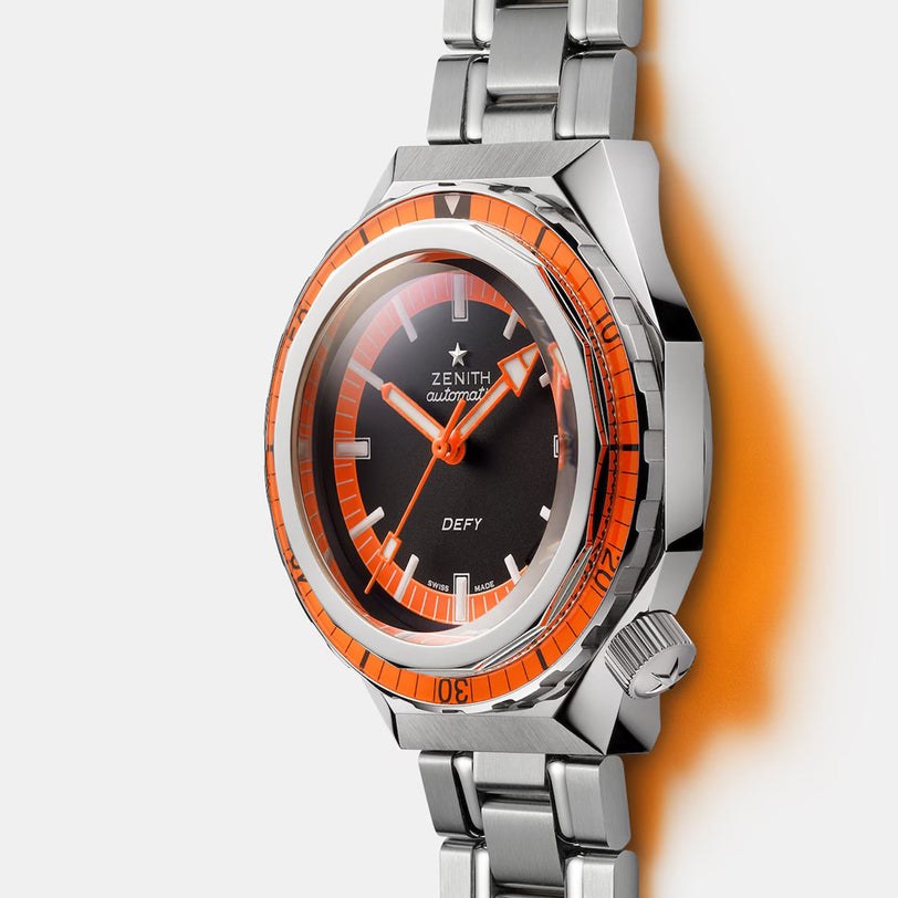 A Zenith Defy Revival diver’s watch with a stainless steel band, black dial, and an orange unidirectional rotating bezel is photographed on a white background.
