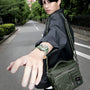 A person in a black jacket and green bag extends their arm, displaying the Zenith Limited Edition Pilot Big Date Flyback Porter with a khaki-green ceramic case, on an empty street lined with greenery.