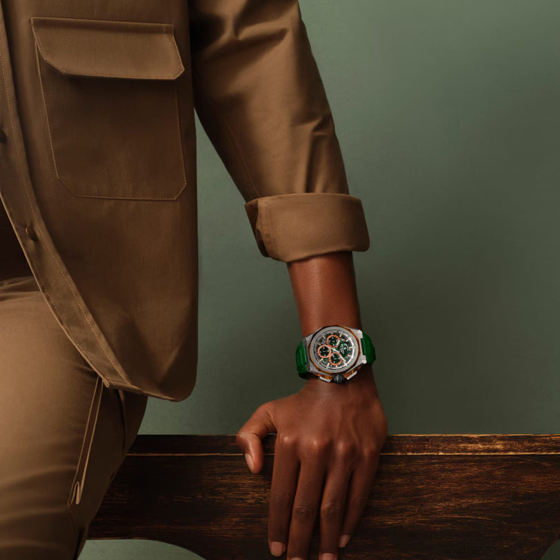 A person in a brown suit leans on a wooden surface, displaying a Zenith Defy Extreme Jungle Edition chronograph with a green strap and intricate dial, set against a plain green background.