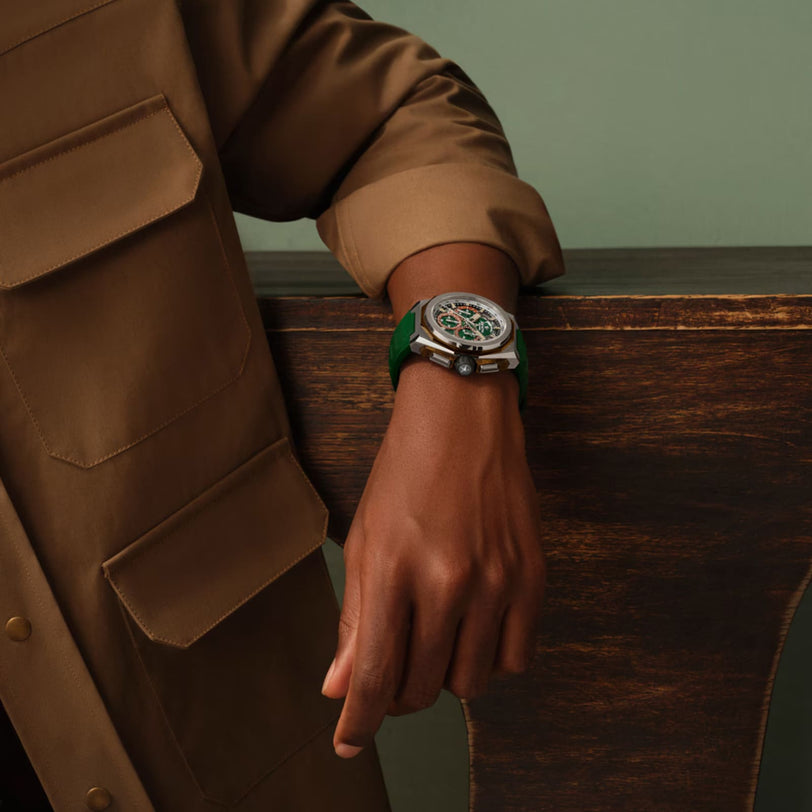 A person in a tan button-up jacket rests their arm on a wooden surface, showcasing the Zenith Defy Extreme Jungle Edition watch, featuring a green strap and an intricate multicolored dial.