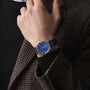 A person in a brown and black houndstooth jacket and black shirt showcases the Zenith Limited Edition G.F.J. Calibre 135 160th Anniversary Edition wristwatch with a platinum round case, blue dial, and blue strap.