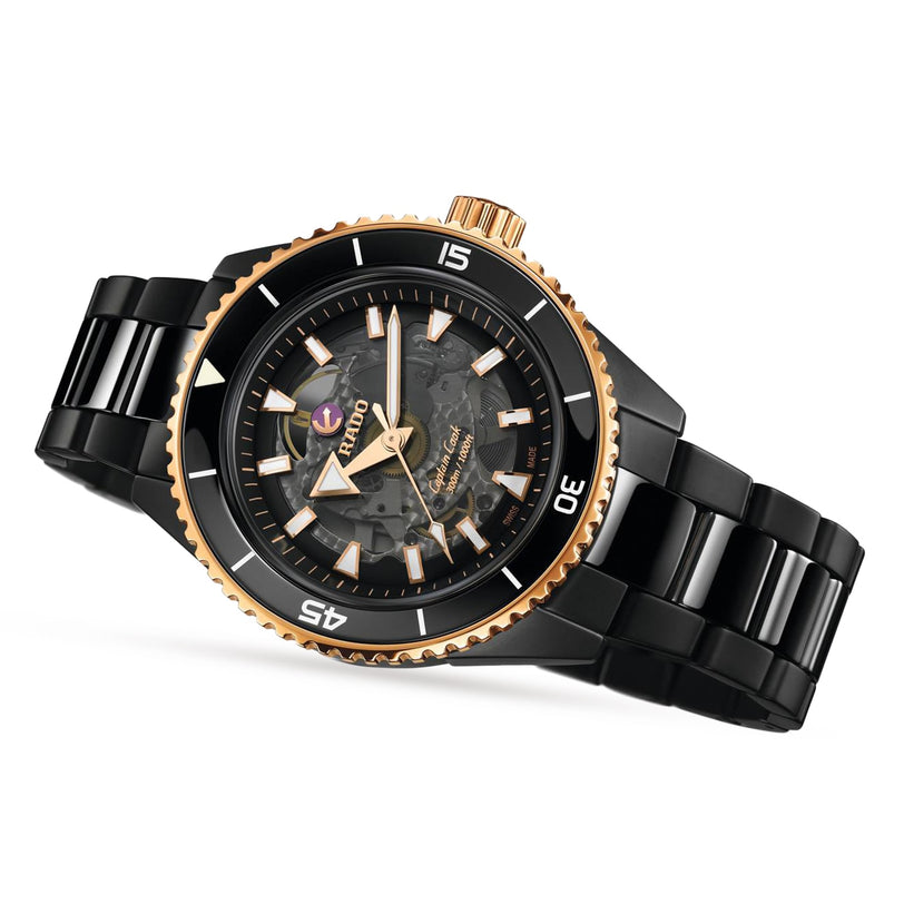 The Rado Captain Cook High-Tech Ceramic wristwatch features a black ceramic band, gold bezel, black dial with luminous markers, and high-tech ceramic elements, showcased at an angle on a white background.