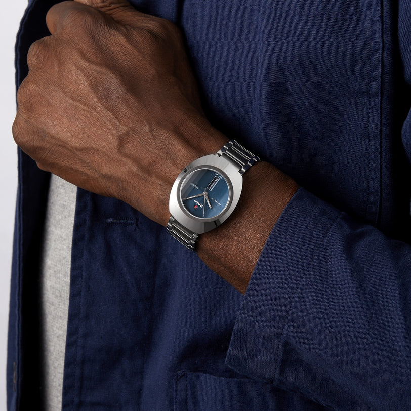 A person wearing the Rado DiaStar Original silver wristwatch with a blue face and sapphire crystal, dressed in a blue jacket over a light gray shirt.