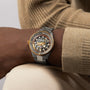 Close-up of a person wearing the Rado Captain Cook High-Tech Ceramic Skeleton wristwatch, featuring a gold and silver design with a skeleton dial, paired with beige and brown attire.