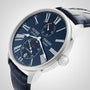 A silver Ulysse Nardin Marine Torpilleur watch with a blue dial, Roman numerals, two subdials, and a blue leather strap is shown angled against a light background.