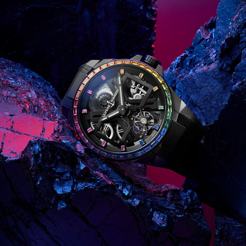 The Ulysse Nardin Blast Tourbillon Rainbow watch, featuring a striking skeleton X dial and black case adorned with vibrant gemstones, dazzles atop dark textured rocks under captivating pink and blue lighting.