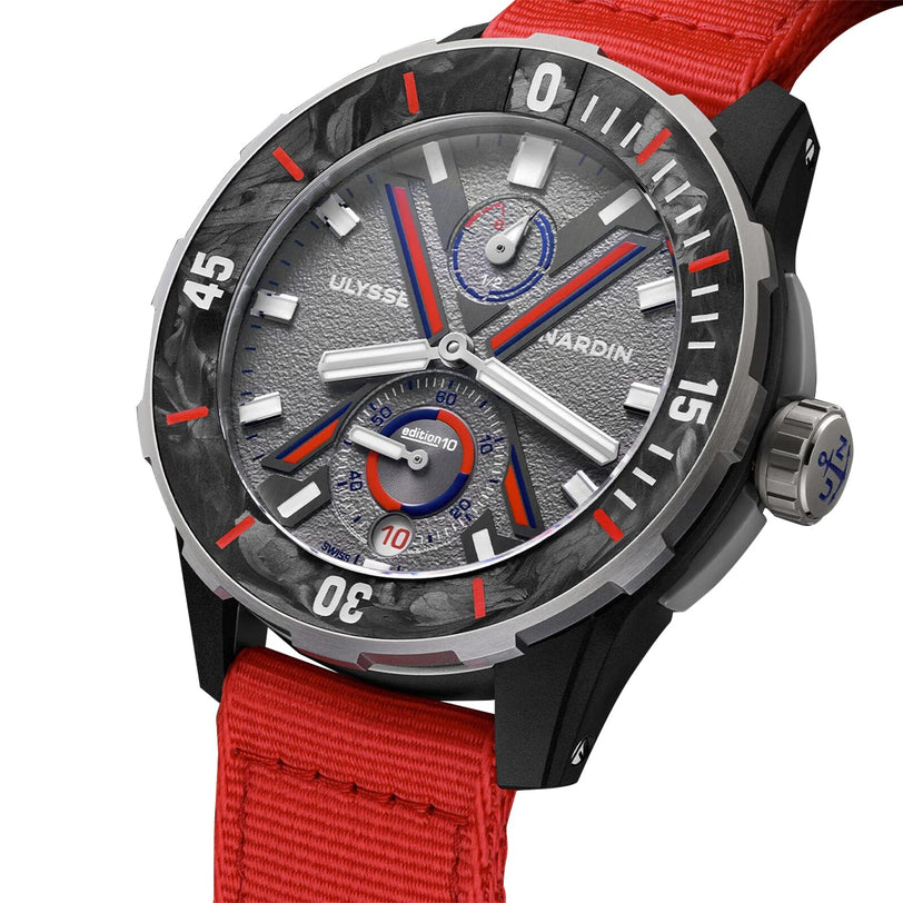 The Ulysse Nardin Diver NET Vendée Globe features a textured gray dial with red and blue accents, subdials, white bezel numerals, and a red fabric strap. Crafted sustainably, it honors round-the-world sailing; the crown sports a blue anchor logo.