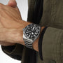 A person wearing the TUDOR Pelagos, a divers tool watch with a black dial and bezel on a titanium bracelet, dressed in a green jacket.