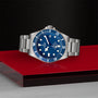 The TUDOR Pelagos, a stainless steel diving watch with a blue dial and bezel, is displayed on a red and black geometric surface showing the time 10:11.