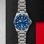 A TUDOR Pelagos wristwatch with a blue bezel and dial, silver hour markers, and date display rests on a textured black and metallic surface with a red accent.