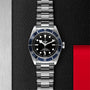 A TUDOR Black Bay wristwatch with a stainless steel bracelet and black dial is set against a textured black background with silver and red accents.