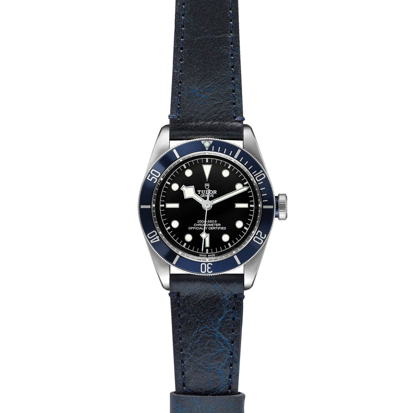 A TUDOR Black Bay wristwatch, with a black dial, white hour markers, blue bezel, and blue leather strap, is displayed on a white background—a striking modern diver’s watch.